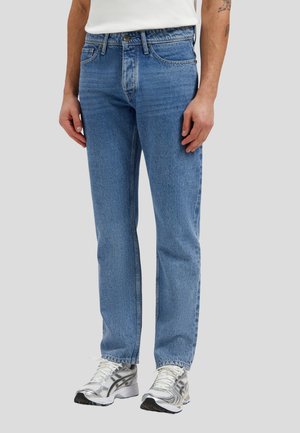 Male wearing straight-leg blue denim jeans and white sneakers, standing against a plain light gray background.