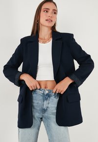 Sötétkék blézer, bemetszett gallérral és két elülső zsebbel. Fehér bordázott crop top fölött viselve, világoskék farmernadrággal párosítva.
