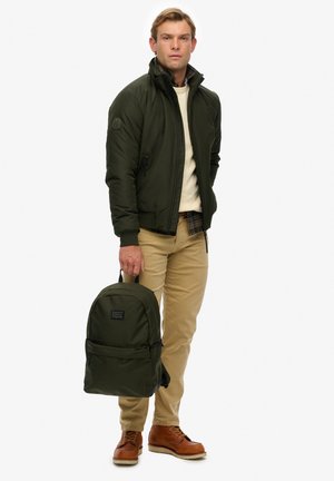 Hombre con chaqueta verde oscuro y pantalones beige, sosteniendo una mochila verde oscuro, de pie frente a un fondo blanco.