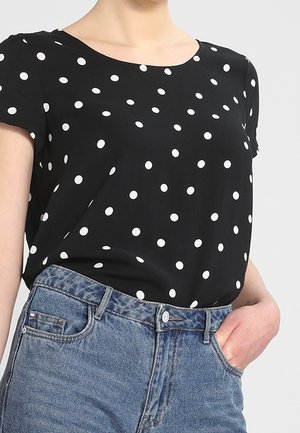 Kobieta w czarnej koszulce z krótkim rękawem w białe grochy, włożonej do niebieskich dżinsów z wysokim stanem, ręka w kieszeni.