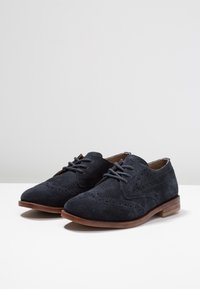 Scarpe in suede blu scuro con lacci, dettagli brogue, punta arrotondata e tacco di legno basso e impilato. Superficie texturizzata e design classico.