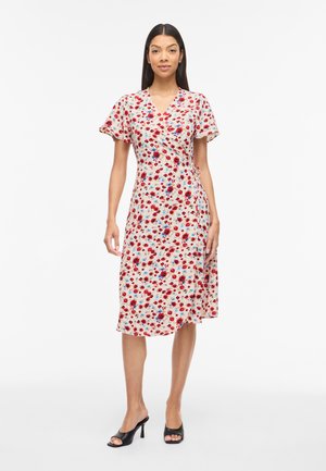 Kobieta w kremowej sukience kopertowej do kolan z czerwono-niebieskim kwiatowym wzorem i zwiewnymi rękawami, w połączeniu z czarnymi sandałami na obcasie z odkrytymi palcami.