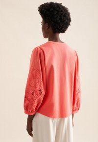 Koralowa bawełniana bluzka z puffowanymi rękawami, ozdobiona haftami kwiatowymi i ażurowymi wycięciami. Okrągły dół, miękka tekstura i luźny krój.