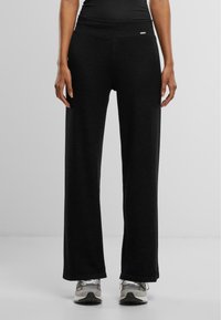 Schwarze weite Hosen mit hohem Bund, aus einem weichen, dehnbaren Stoff gefertigt. Besitzt ein kleines Logo-Tag am Bund.