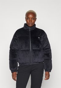 Giubbotto puffy nero in peluche con colletto alto, zip frontale completa e polsini elasticizzati. Presenta un piccolo logo triangolare sul petto.