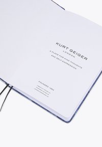 Carnet Kurt Geiger avec une page intérieure blanche, une couverture texturée bleue et un marque-page en ruban noir. Présente un logo et un slogan imprimés.