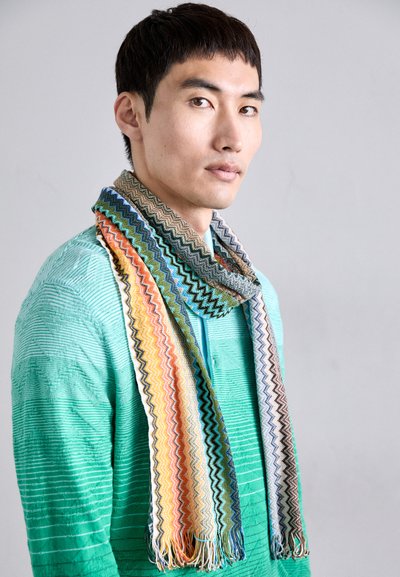 Cachecol com padrão zigzag multicolorido e bordas franjas, feito de um tecido suave. Usado sobre uma blusa verde texturizada, exibindo várias tonalidades.