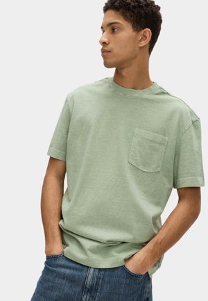 Ung mand med kort krøllet hår iført en lys grøn T-shirt med korte ærmer og brystlomme samt blå jeans, hænderne i lommerne, kigger til siden.