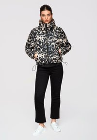 Femme debout les mains dans les poches, portant une veste imprimée léopard noir et blanc, un pantalon court noir et des baskets blanches, sur un fond gris.