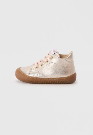 Sneaker basse dorée métallisée avec des lacets rose clair, bout rond, upper en cuir texturé, et semelle en caoutchouc, mettant en valeur des détails de couture et des œillets.