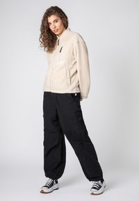 Beige Fleece-Jacke mit Reißverschluss vorne, zwei Seitentaschen und einer Brusttasche. Kombiniert mit schwarzen Cargo-Hosen und schwarzen-weißen Turnschuhen.