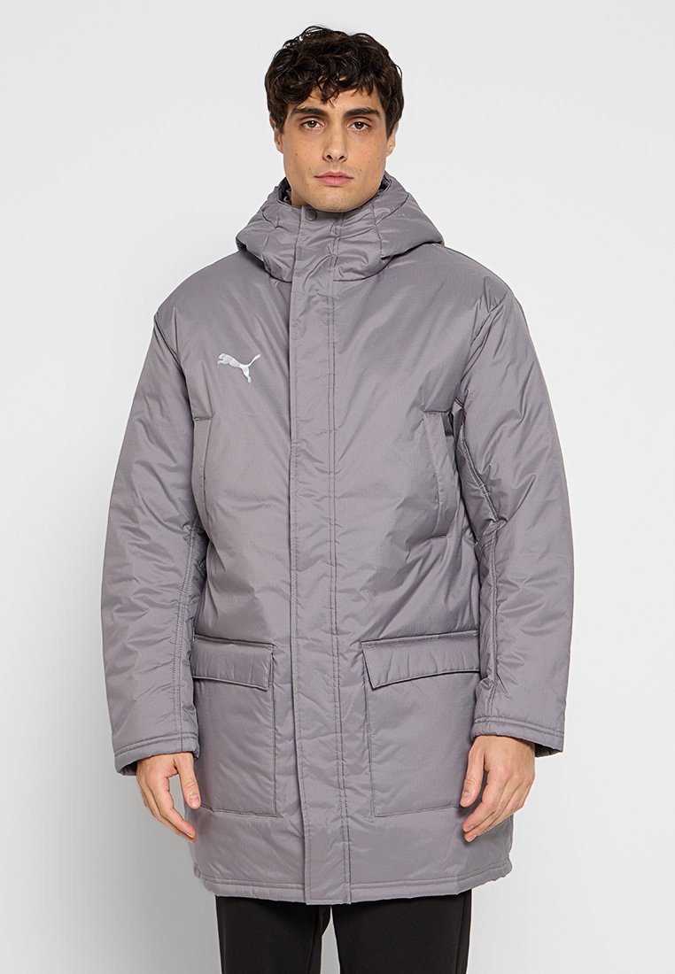 Hombre vestido con una chaqueta acolchada gris con capucha, con bolsillos frontales y un logo de Puma en el pecho, de pie contra un fondo sencillo.