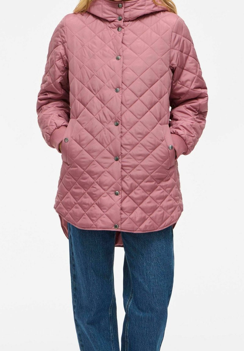 Femme portant une veste matelassée rose avec des boutons-pression et des poches, associée à un jean en denim bleu, debout les mains dans les poches.