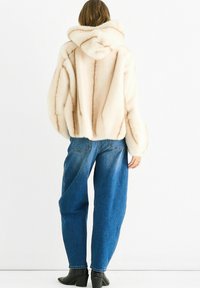 Person wearing a cream fur hooded jacket, loose blue jeans, and black heeled ankle boots, standing against a white background.