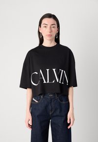 Černé krátké tričko s výrazným bílým nápisem "CALVIN". Vyrobeno z měkké tkaniny s kr�átkými rukávy a volným střihem. V kombinaci s tmavými džínami.