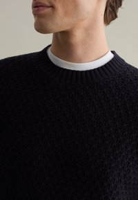 Marine gebreide pullover met een textuurpatroon, een geribde halslijn en een gelaagd wit kraagdetail eronder.