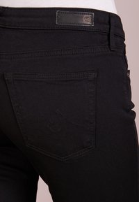 Černé džínové kalhoty s detailním pohledem na zadní kapsu, která má zakřivený steh a koženou nášivku značky nad pasem.