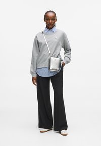 Kobieta w szarej bluzie nosząca niebieską koszulę w kratę, czarne spodnie szerokie, białe sneakersy i srebrną torbę na ramię, stoi na tle białego tła.