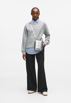 Kobieta w szarej bluzie nosząca niebieską koszulę w kratę, czarne spodnie szerokie, białe sneakersy i srebrną torbę na ramię, stoi na tle białego tła.