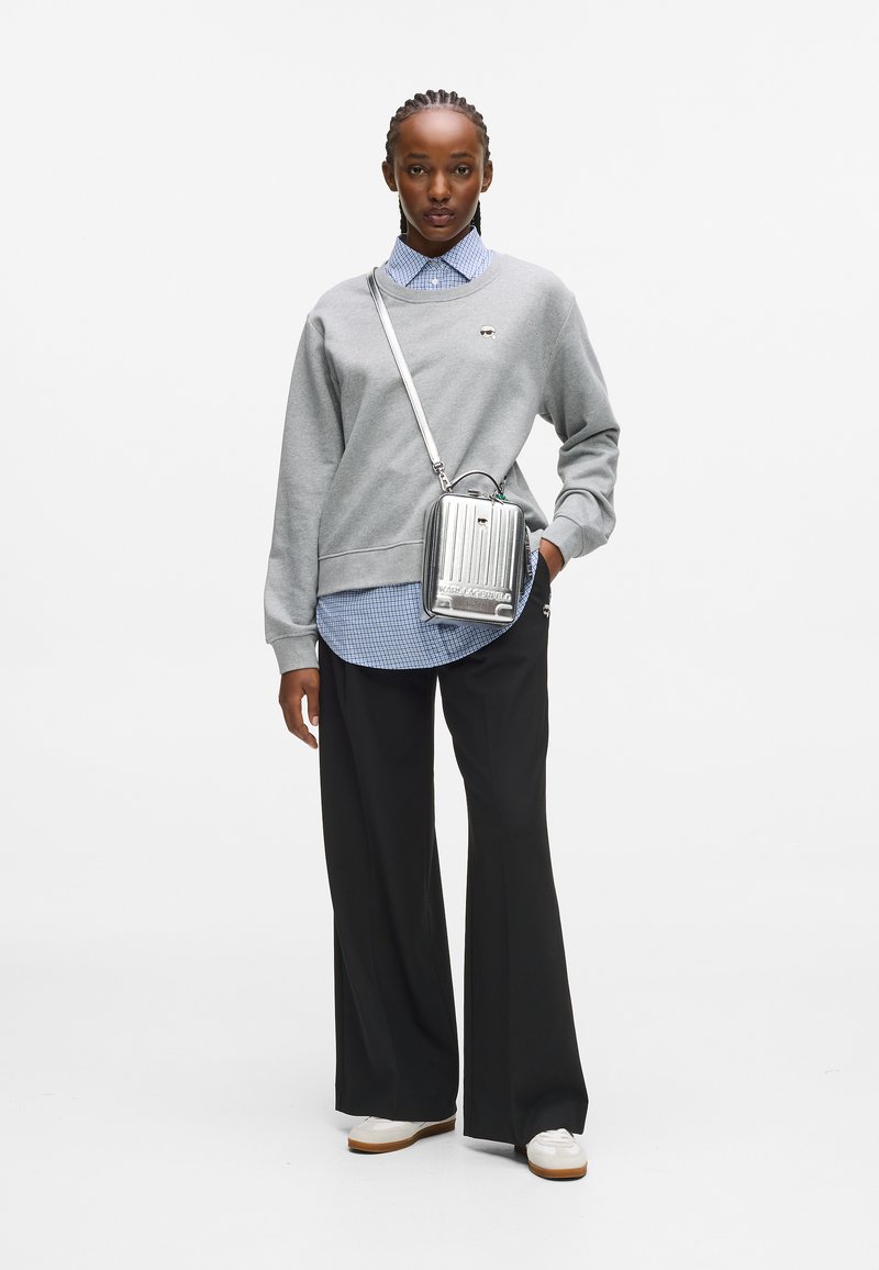 Kobieta w szarej bluzie nosząca niebieską koszulę w kratę, czarne spodnie szerokie, białe sneakersy i srebrną torbę na ramię, stoi na tle białego tła.