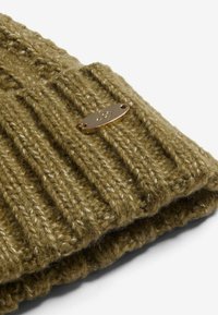Olivově zelená pletená čepice s ribovým vzorem a malou zlatou logovou destičkou. Měkká, silná příze s viditelnými švy.