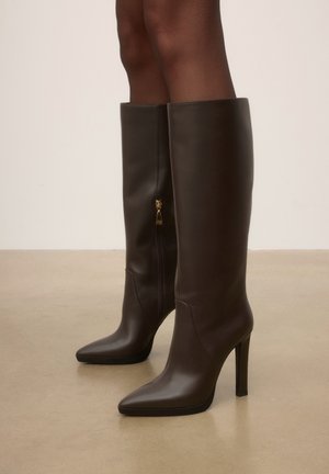 Bottes en cuir marron foncé montantes jusqu'aux genoux avec des bouts pointus et des talons blocs hauts, portées sur une peau foncée.