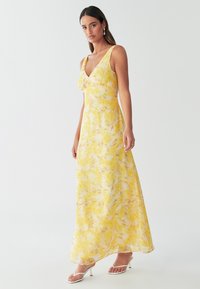 Yellow floral maxi dress with a v-neckline, wide straps, and flowy fabric. Features a light, airy texture and a slight gathering at the bust.