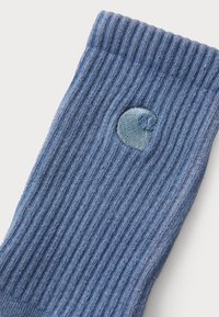 Ribbet blå sok med et cirkulært broderet logo i en lysere blå nuance nær den øverste del. Lavet af et blødt, struktureret materiale.