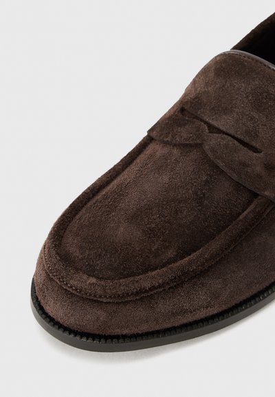 Brūni zamšaini loafers ar noapaļotu purngalu, kurus rotā šuvums un pacelta lente pāri augšai. Gumijas zole ir ar tekstūru, lai nodrošinātu labāku saķeri.