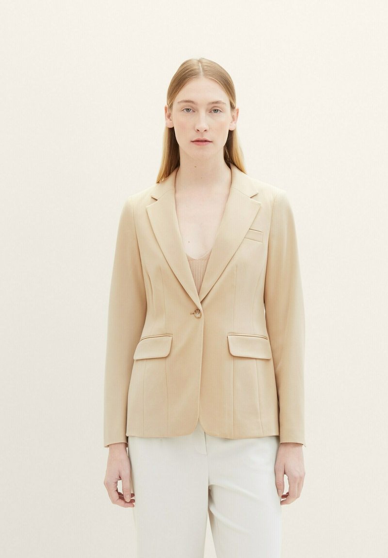 Donna che indossa un blazer beige con un solo bottone sopra una maglietta chiara, abbinato a pantaloni bianchi, in piedi davanti a uno sfondo chiaro e uniforme.