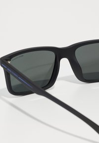 Ochelari de soare rectangulari negri cu lentile gri, finisaj mat și accent albastru pe braț. Design geometric simplu, fără modele suplimentare.