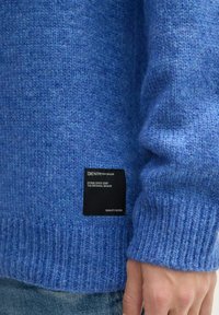 Blaue Strickjacke aus weichem Material, mit gerippten Bündchen und einem schwarzen Markenetikett mit weißem Text in der Nähe des Saums.