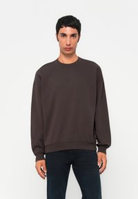 Young man standing, wearing a loose dark brown sweatshirt and dark jeans, against a plain white background.