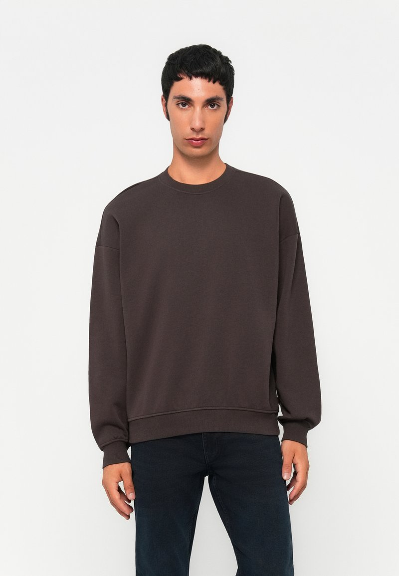 Young man standing, wearing a loose dark brown sweatshirt and dark jeans, against a plain white background.