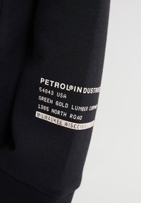 Sudadera negra con texto impreso en blanco en la manga, que incluye "PETROL INDUSTRIES", detalles de dirección y ubicación en Wisconsin.