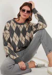 Sivi in krem pulover z argyle vzorcem in sproščenim krojem, v kombinaciji s svetlo sivimi kavbojkami in belimi supergami. Očala velikega kroga so na obrazu.