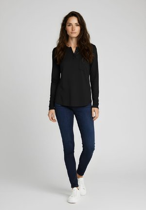 Mujer con cabello largo y castaño, vestida con una camiseta negra de manga larga, jeans ajustados de mezclilla azul oscuro y zapatillas blancas, posando frente a un fondo liso.