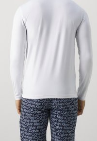 Hombre vestido con una camisa blanca de manga larga y pantalones azul marino con el logotipo blanco repetido de "Calvin Klein", visto desde atrás sobre un fondo liso.