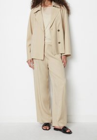 Beżowa dwurzędowa marynarka z klapami, w połączeniu z pasującymi szerokimi spodniami. Noszona z prostymi czarnymi sandałami na płaskiej podeszwie.