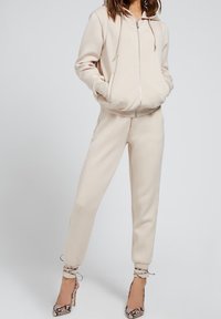 Sudadera beige con cremallera, cordón y bolsillos laterales, combinada con pantalones de chándal ajustados a juego y zapatos de tacón con estampado de serpiente.