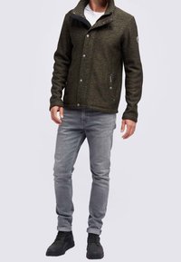 Uomo che indossa una giacca buttonata verde scuro, jeans grigi chiari, camicia bianca e scarpe nere alte, in piedi contro uno sfondo semplice.