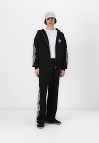 Svart hettegenser med sølvfarget grafisk design på ermene, kombinert med svarte joggebukser. Personen har på seg en hvit t-skjorte og en lys grå caps.
