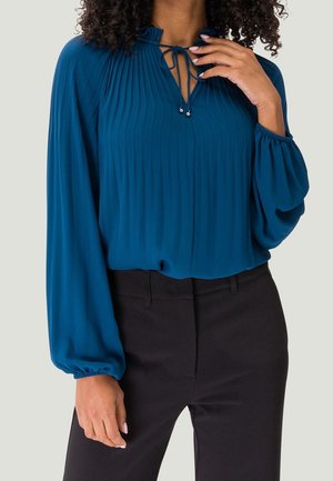 Vrouw die een tealblauwe geplooide blouse draagt met lange, opgeblazen mouwen en zwarte, nauwsluitende broek, met een hand die de halskoorden met metalen kralen aanraakt.