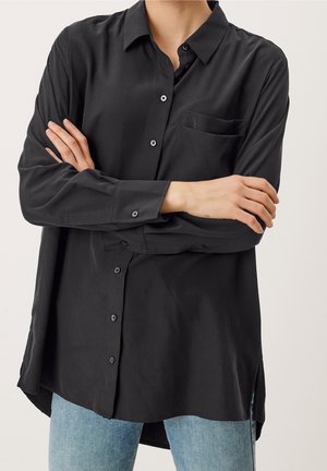 Persona vistiendo una camisa negra holgada de botones con un bolsillo en el pecho y jeans azul claro, con los brazos cruzados sobre el pecho.