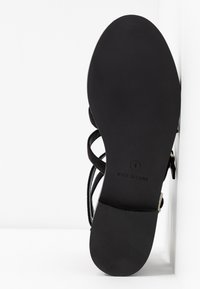 Musta sandaali, jossa on littee pohja ja ohuet remmit. Pohjassa on teksturoitu pinta ja siinä on koko- ja "Valmistettu Kiinassa" -merkinnät.