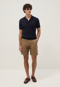 Hombre con gafas de sol, polo negro de manga corta, pantalones cortos beige y zapatos de cuero marrón, de pie con las manos en los bolsillos.