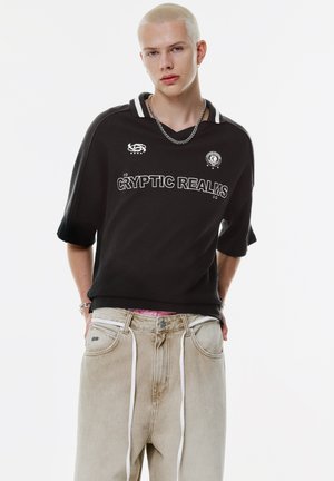 Persona bionda che indossa una maglietta nera "Cryptic Realms", pantaloni beige larghi con lacci bianchi, collana di catena d'argento e piccolo orecchino a cerchio.
