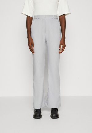 Pantalons larges gris clair en tissu texturé. Dotés d'une taille avec passants de ceinture et de plis à l'avant, associés à des chaussures noires.