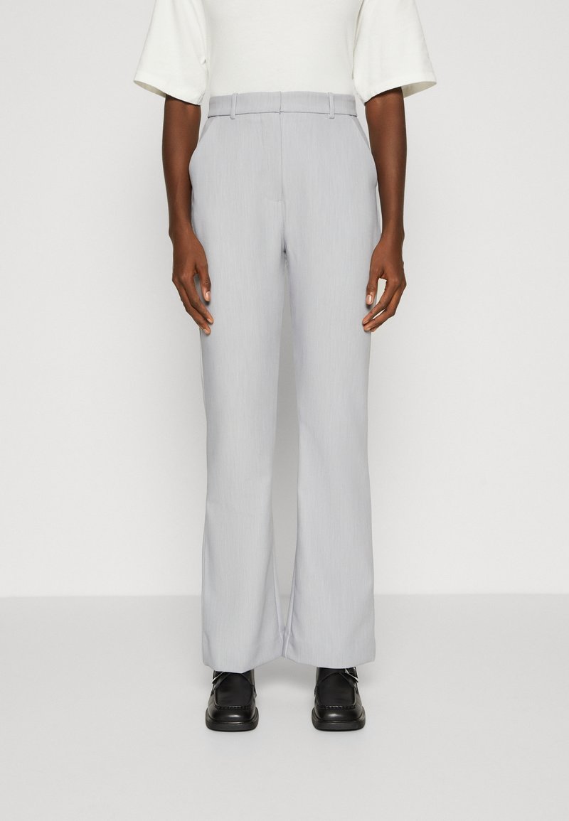 Světlé šedé kalhoty širokého střihu z texturované látky. Mají pas s poutky na pásek a přední sklady, v kombinaci s černými botami.