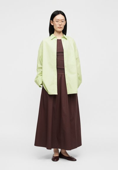 Femme portant une chemise boutonnée oversize vert clair sur un top marron sans manches et une longue jupe avec des ballerines marron, debout sur un fond gris.